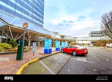 Red electric vehicle charging at a public station in downtown Portland, with multiple EV chargers lined along the sidewalk and city buildings in the background.