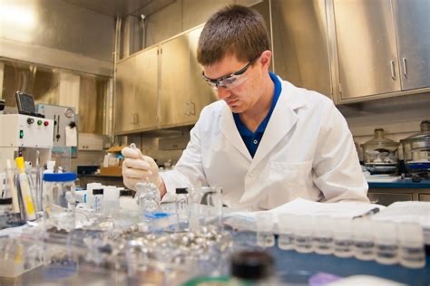 U.S. Army researcher in a lab coat and safety glasses works with battery cells and test equipment in an Army Research Laboratory bench setting.