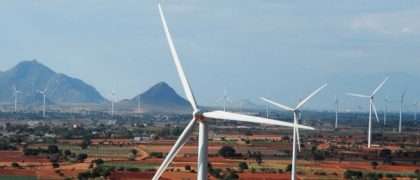 Multiple wind turbines owned by Gamesa generating electricity in a dry landscape with hills under a partly cloudy sky
